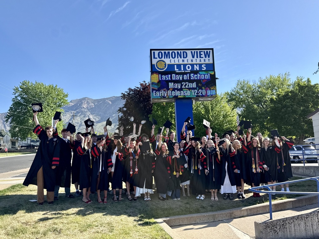 The graduating class standing outside in front of the Lomond View Elementary sign, cheering and tossing their caps into the air with mountains in the background.