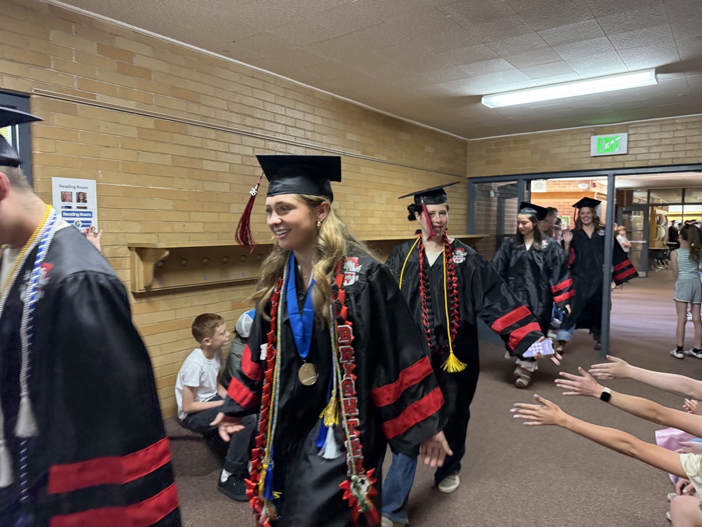 A high school graduate in a black and red cap and gown smiles while walking down an elementary school hallway, high-fiving students.