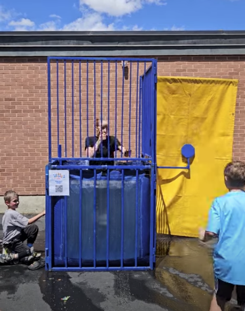 A series of photos showing Dr. Watson, the principal of Lomond View Elementary, participating in a dunk tank event on a sunny day. She is wearing a black shirt and sitting on the collapsible platform above a large blue water tank. One photo captures the moment a student hits the target, and subsequent photos show Dr. Watson submerged in the water and then emerging with a big smile while students cheer her on.