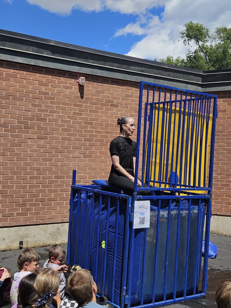 A series of photos showing Dr. Watson, the principal of Lomond View Elementary, participating in a dunk tank event on a sunny day. She is wearing a black shirt and sitting on the collapsible platform above a large blue water tank. One photo captures the moment a student hits the target, and subsequent photos show Dr. Watson submerged in the water and then emerging with a big smile while students cheer her on.