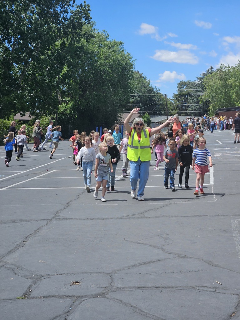 An energetic scene on the school blacktop where a staff member in a bright neon yellow safety vest and blue overalls leads a large group of students in a dance party. She has her arms raised and a wide smile, and the students are following along enthusiastically.