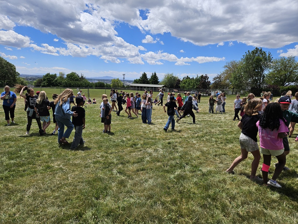 A wide shot of a grassy field at Lomond View Elementary under a blue sky with white clouds. Dozens of students are scattered across the grass participating in water relay games, while teachers in the background supervise the activity.