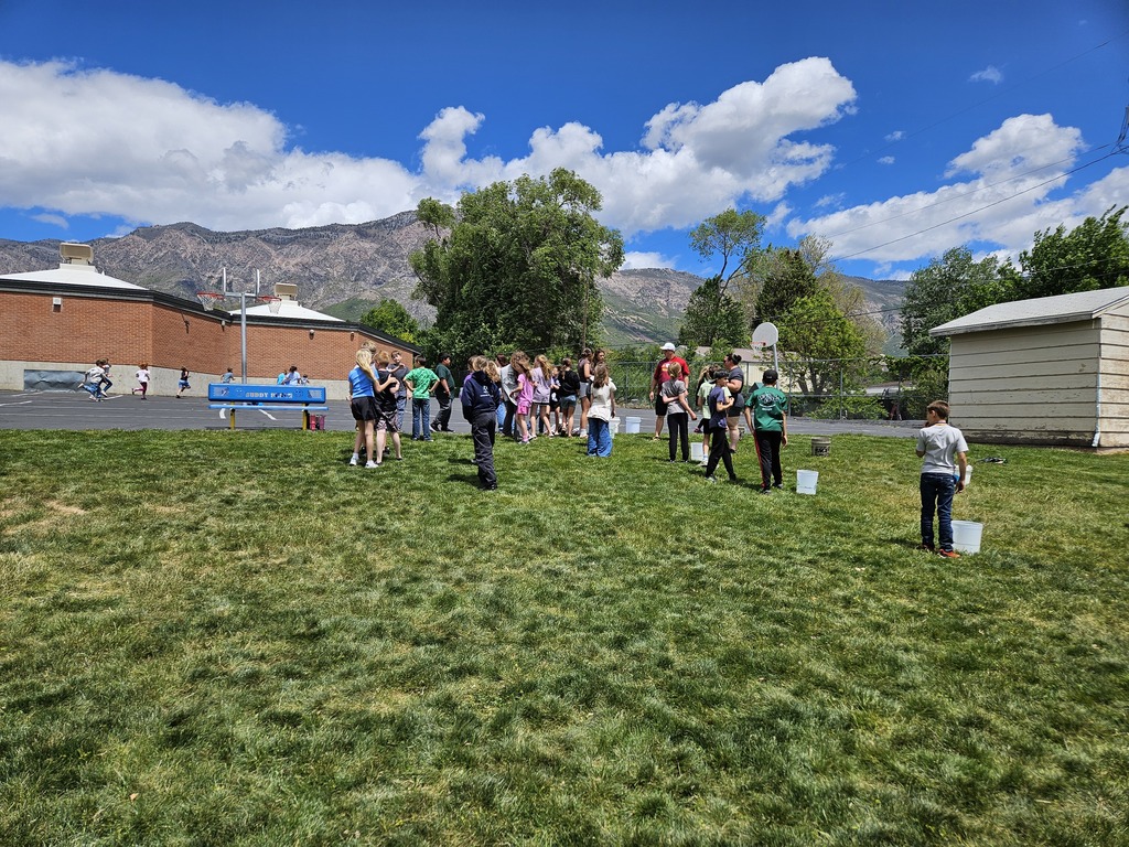  Students stand in a line on a grassy field with white buckets at their feet, preparing for a water relay race. The majestic Wasatch mountains are visible in the background behind the school building and trees.