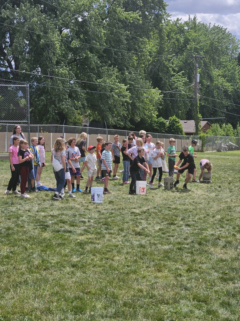 A close-up of students on the field during a water game. Two students are focused on transferring water or an object between buckets while their classmates watch and cheer from the line.