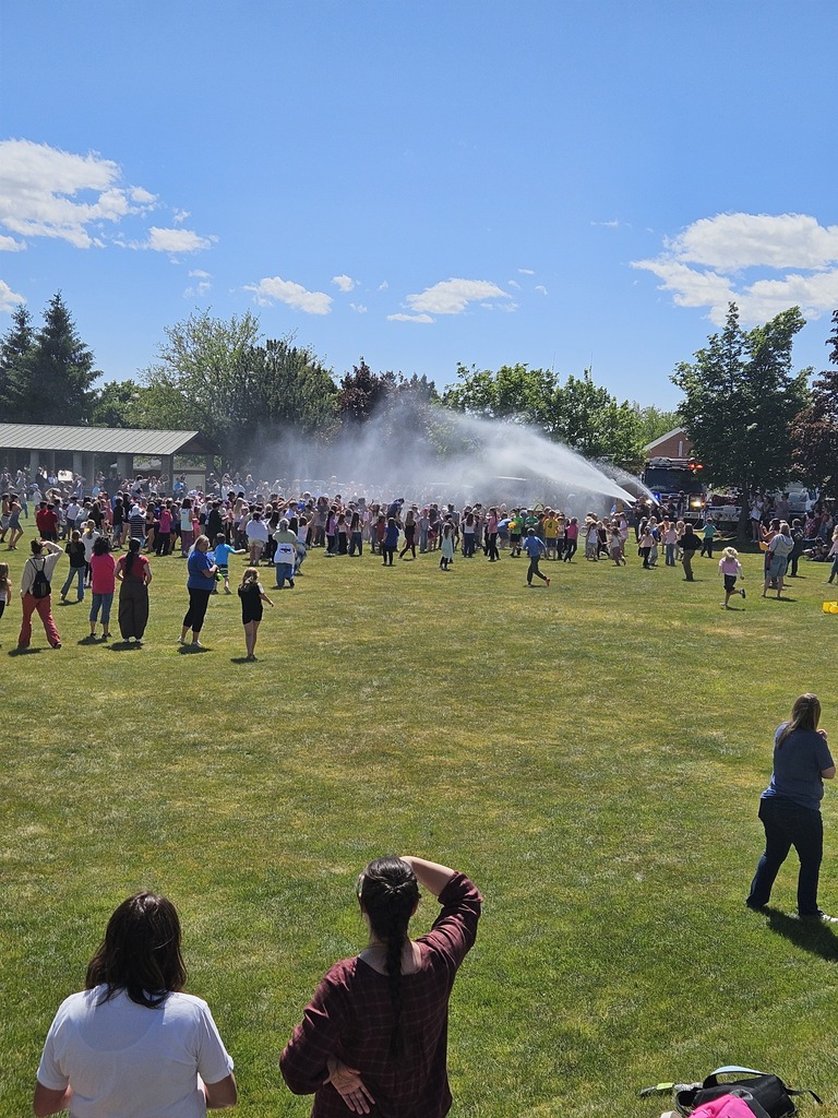 A wide, sunny shot of the Lomond View Elementary field where a Pleasant View fire engine is parked at the edge of the grass. A high-pressure water hose from the truck is spraying a massive, shimmering arc of water over a large crowd of cheering students. Kids are running and dancing through the mist under a bright blue sky, while several teachers and parents watch the excitement from the foreground.