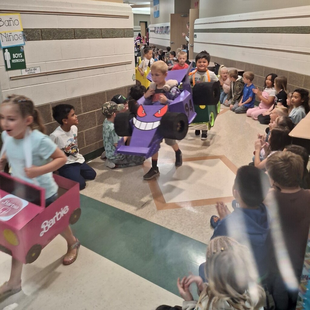 Kids in cardboard cars walking in the hall lined with students 