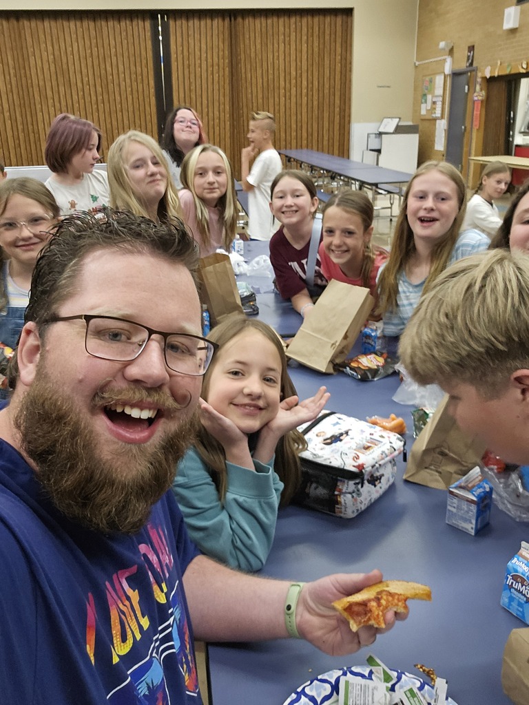 A smiling teacher with a beard and glasses taking a selfie with a group of elementary students sitting at a cafeteria table during lunch. He is holding a piece of pizza, and the kids are grinning and posing for the camera.