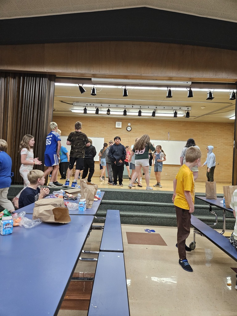 A wide shot of an elementary school cafeteria/gym. A group of students are dancing and playing on a raised wooden stage under stage lights, while other students sit at blue cafeteria tables in the foreground.