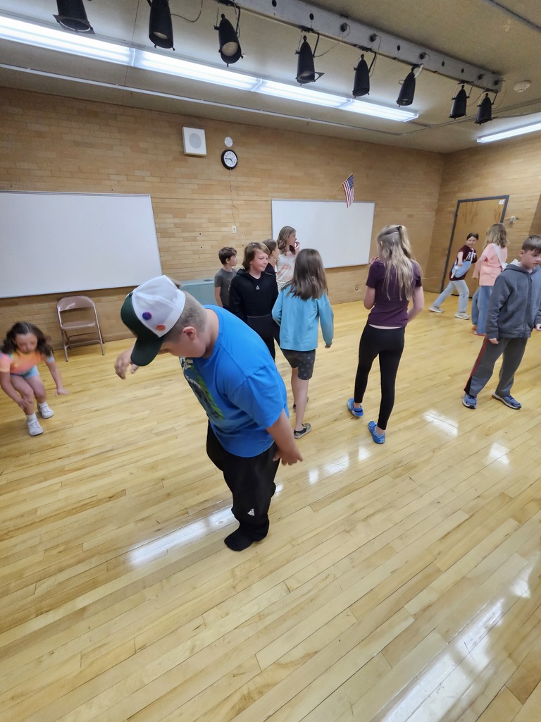 A close-up view of the stage where several elementary students are mid-dance move. One boy in a blue shirt and hat is leaning forward, and others are scattered around the wooden floor enjoying an impromptu dance party.