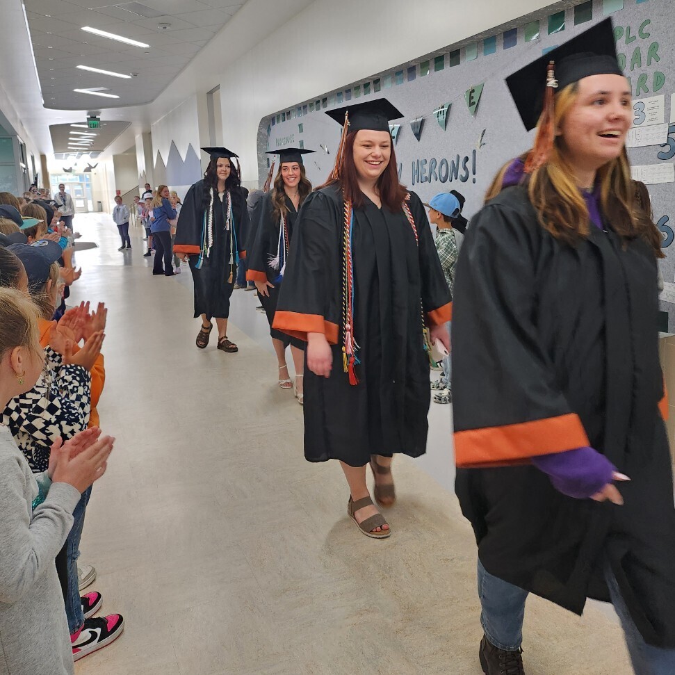 A group of female high school graduates smiling as they walk down a school hallway lined with cheering elementary students. The graduates wear black caps and gowns with orange trim, and several wear multicolored graduation cords. In the background on the right, a bulletin board displays the text "HAVEN BAY HERONS!" along with "PLC CARD BOARD" and numbers listed vertically.