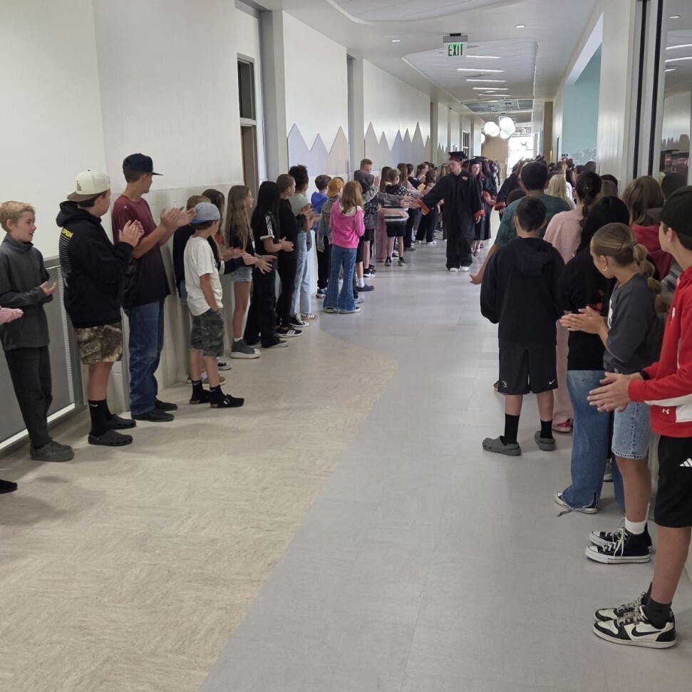 A long, brightly lit school hallway filled with elementary-aged students lining both sides of the corridor, clapping and cheering. Down the center of the hallway, high school graduates wearing black caps and gowns with orange accents walk through, high-fiving the younger students. An illuminated green "EXIT" sign hangs from the ceiling in the distance.