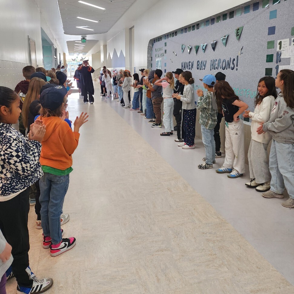 A wide shot of a modern school hallway where elementary students line the walls, clapping in anticipation. In the far distance, a graduate in a black cap and gown begins walking down the center of the hall. On the right wall, a large bulletin board features the text "HAVEN BAY HERONS!" in bold letters, decorated with hanging pennants and additional words including "RESPECTFUL".