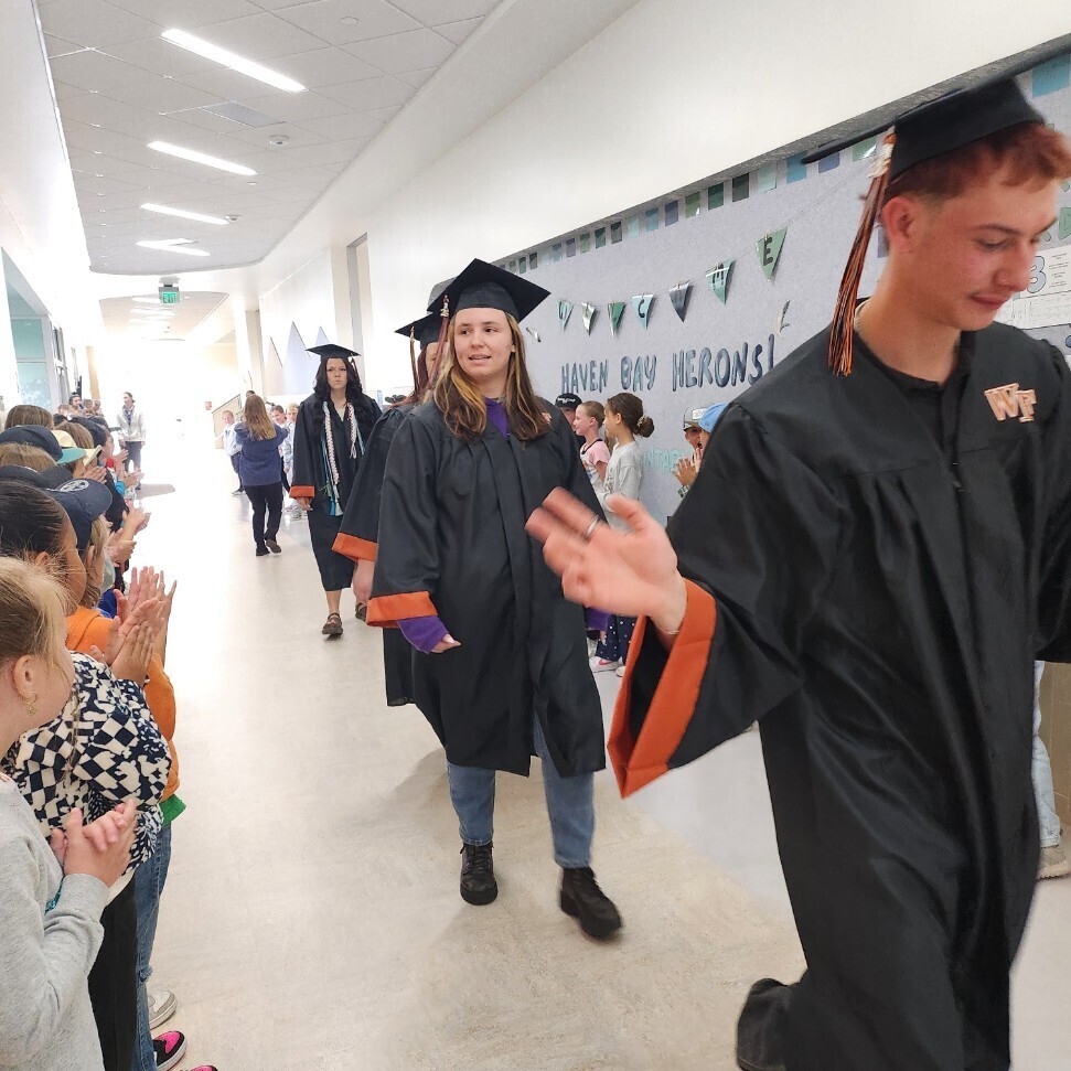 A close-up, eye-level shot of high school graduates walking down a school hallway while elementary students line the left side, clapping. In the foreground, a male graduate in a black gown with orange cuffs and a "WF" monogram on the chest smiles and reaches out his hand to high-five the kids. Behind him, a female graduate in a cap and gown walks forward. The bulletin board on the right wall reads "HAVEN BAY HERONS!".