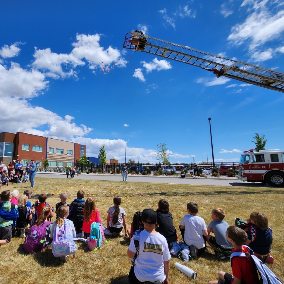 A large group of children is kneeling on a grassy field in front of a modern school building. They are facing away from the camera, looking towards a huge red fire engine on the right. A long, extended ladder from the truck is high in the sky. It appears that objects are being thrown or dropped from a basket at the end of the ladder. An adult is standing in front of the kids, gesturing towards the ladder. The sky is partially cloudy. The photo captures a moment of excited anticipation.