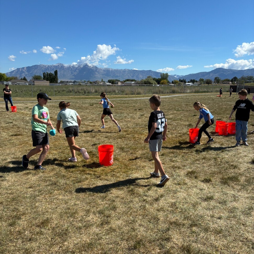 A group of children is playing an outdoor game on a grassy field on a clear, sunny day. The game involves orange buckets placed on the grass and blue water sponges. A boy in a green shirt is about to dip his sponge in a bucket. To the right, another boy is running, and a girl is near a stack of orange buckets. The background shows mountains with snow-capped peaks. An adult is visible in the far-off distance. The scene is bright and full of action.