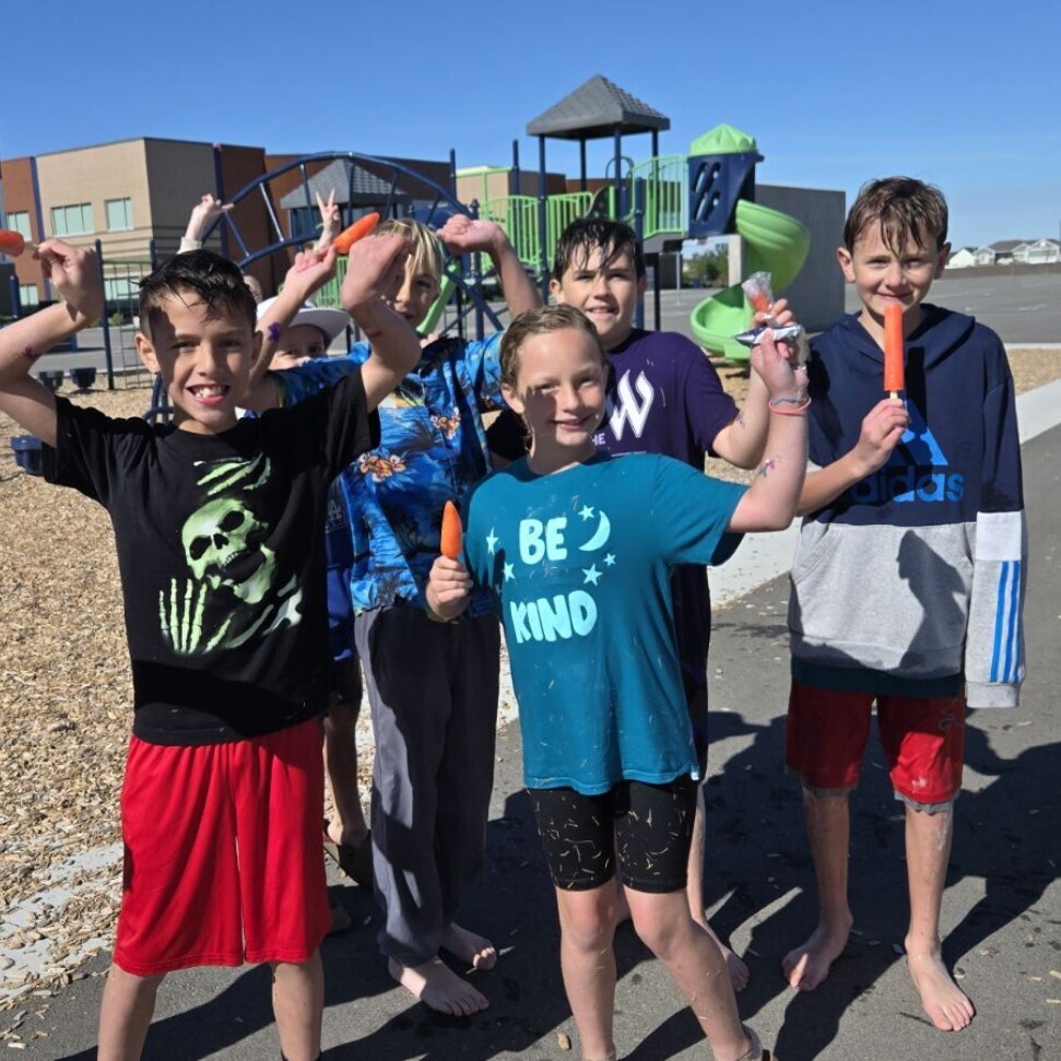 A group of eight elementary school-aged kids is posing happily on a playground on a sunny day. They are standing near a green and blue jungle gym, and one boy is on a red slide. All children are holding orange and red popsicles, smiling at the camera. Two boys are barefoot on the asphalt track, while others wear shoes. Their hair and clothes appear slightly wet. The sky is bright blue. The photo has a cheerful and summery feeling.