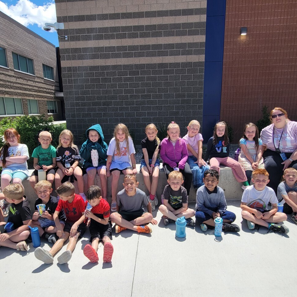A class photo of fifteen young students, boys and girls, and their female teacher, posing on a concrete path outside their school. All students are sitting or kneeling, holding blue water bottles, looking at the camera. Their teacher is on the right, kneeling and smiling. The students' hair looks wet, indicating they have been participating in water activities. The modern, two-story school building is behind them, with brick and siding. A security camera is visible on the building. The photo is well-lit on a bright, sunny day.