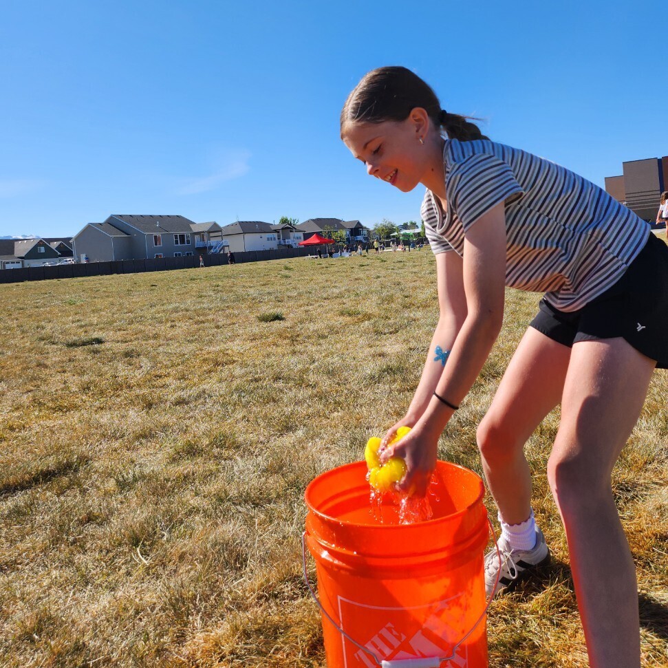 A pre-teen girl is smiling as she dunks a yellow sponge into an orange bucket filled with water. She is on a dry, grassy field on a bright, sunny day. The girl is in motion, as she is bringing her hands up from the bucket. A butterfly tattoo is visible on her right forearm. The bucket has the Home Depot logo on it. In the distance, a suburban neighborhood is spread out across a flat landscape under a clear blue sky. The photo is in focus and full of life.
