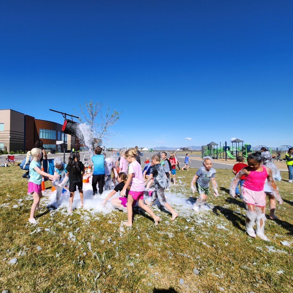 A group of fifteen barefoot children is playing in a large mound of white foam on a grassy lawn. They are clustered in the foam, which covers them and the ground. To the left, a large black bucket is attached to a pole, appearing to be the foam dispenser. Adults are watching in the background. A modern school building, a white pickup truck, and a distant playground are also visible. The sky is clear, dark blue. The scene is full of fun and excitement.