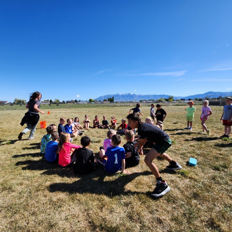A group of about twenty children is participating in an organized activity on a large, grassy field on a clear, sunny day. Most of them are sitting in a circle on the ground, while two adults and four other children are standing outside the circle. The girl in the foreground is in mid-stride, running, likely the person who is "it" in the game. In the distance, a mountain range is clearly visible. The overall mood is active and engaged.