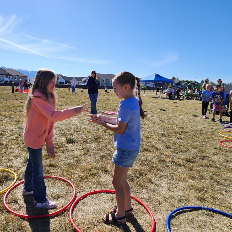 A group of school-aged children is playing "rock paper scissors" while participating in a hopscotch game with multi-colored hula hoops. The game is spread across a large, grassy field on a clear, bright sunny day. A cluster of kids is waiting their turn in the background. A small blue canopy tent is set up further away. A adult is standing to the right. The mountains can be seen in the distance, and the sun is high and bright, creating long shadows. The vibe is active and happy.