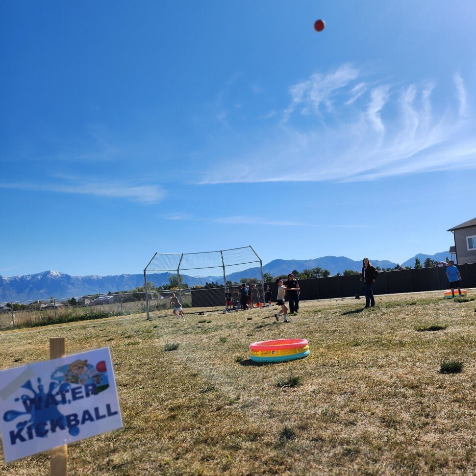 A sign stuck into the ground reads "WATER KICKBALL" with a picture of a kid in a baseball uniform. Beyond the sign, children are playing kickball on a dry, grassy field. A small, multi-colored blow-up wading pool is on the grass near first base. An adult stands near third base. The field is bordered by a tall fence and a chain-link fence. Behind the field, there is a massive mountain range with snow-covered peaks under a vast, cloudless blue sky. The composition captures the wide, open feeling of the landscape.