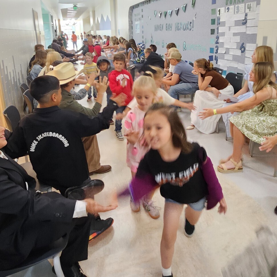 A busy, slightly motion-blurred shot of a bright school hallway where elementary students are running down a high-five line formed by older students and adults sitting in chairs along the walls. A large bulletin board on the right wall has paper decorations and text that reads "HAVEN BAY HERONS!", "BE KIND", "BE SAFE", "BE RESPECTFUL", and "PLC SOAR BOARD" with numbers 1 through 6. A girl in the foreground wears a black shirt reading "WESTFIELD". A boy on the left wears a black jacket reading "NIKE SKATEBOARDING