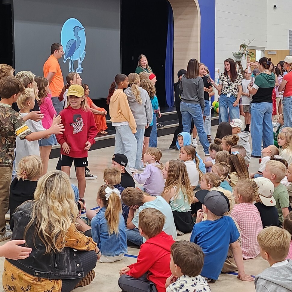 An indoor shot of a school assembly inside a gymnasium or auditorium with high ceilings and colorful acoustic wall panels. A group of young students stands on a low stage next to a large projection screen displaying a blue oval logo with a blue heron bird. Adults with microphones stand near the stage speaking to a large audience of elementary school children sitting cross-legged on the floor.