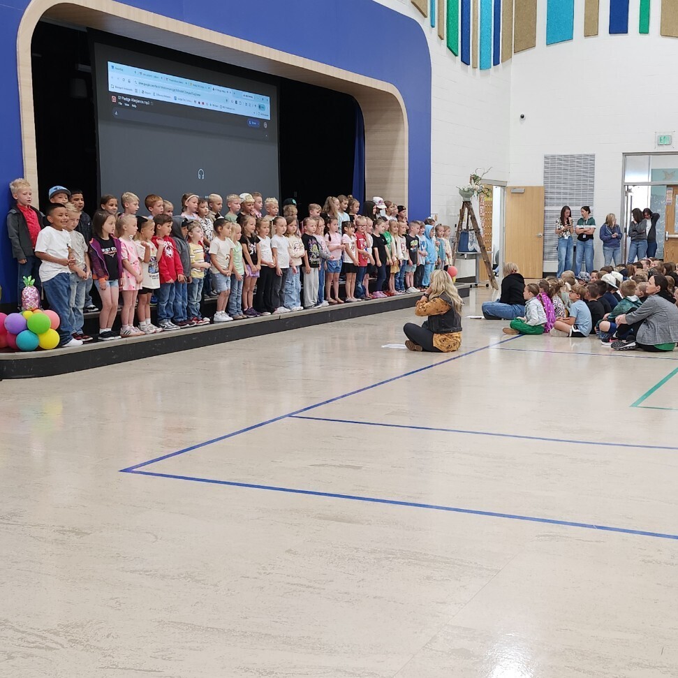 A wide-angle view of a large school assembly inside a spacious gymnasium with bright overhead lighting and a basketball hoop. Hundreds of elementary school students are sitting quietly in rows on the polished floor, facing a stage on the left side where a large choir of children is standing and performing. The back wall features a large, wavy decorative pattern of blue, green, and tan panels.