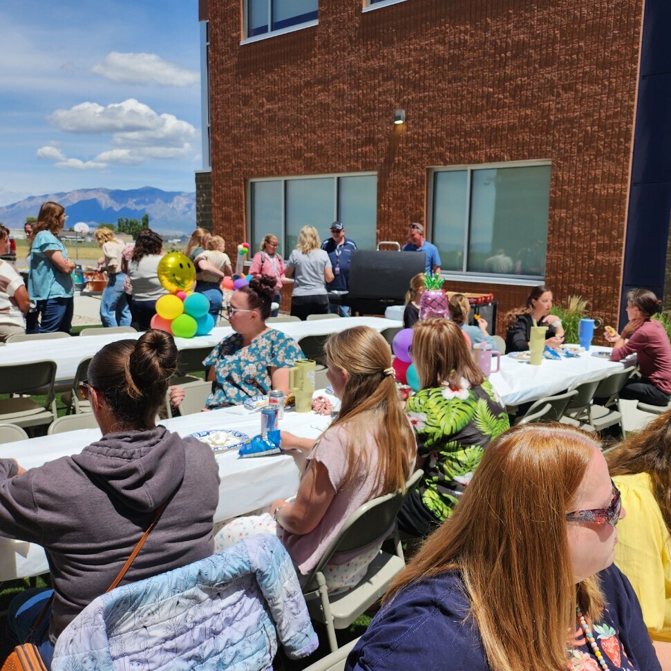 A sunny outdoor luncheon where several women are sitting at long, white cloth-covered folding tables on a grassy area next to a brick school building. They are eating lunch, drinking from cans, and talking. In the background, a grill is set up with people standing around it, and distant blue mountains are visible under a bright sky. Colorful balloon arrangements decoration the tables.