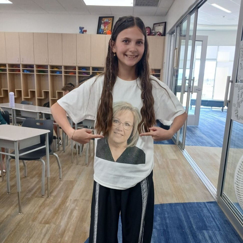 A young girl with long, braided brown hair smiles and stands in the middle of a school classroom with light wood cubbies and desks. She is pointing with both index fingers to a custom white t-shirt she is wearing, which features a large, black-and-white printed photo of a smiling woman with glasses and blonde hair.