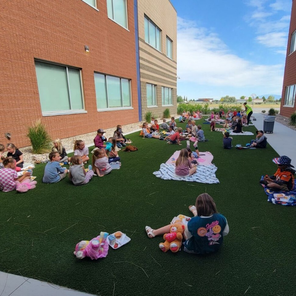 wide outdoor shot of an elementary school courtyard with green artificial turf between two modern brick school buildings under a blue sky with light clouds. Dozens of elementary school students are spaced out across the lawn, sitting on blankets and towels to eat lunch picnic-style. Several stuffed animals and teddy bears are visible next to the children