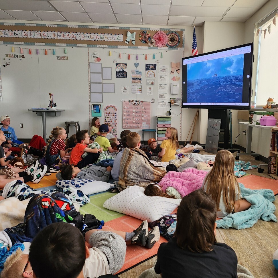 An indoor shot from the side of a gym showing a large group of young elementary students lined up on a stage during an assembly, facing a female instructor who is sitting cross-legged on the floor in front of them. A large audience of students sits on the gymnasium floor watching the presentation. A projection screen on the stage shows a browser tab titled "01 Pledge of Allegiance"