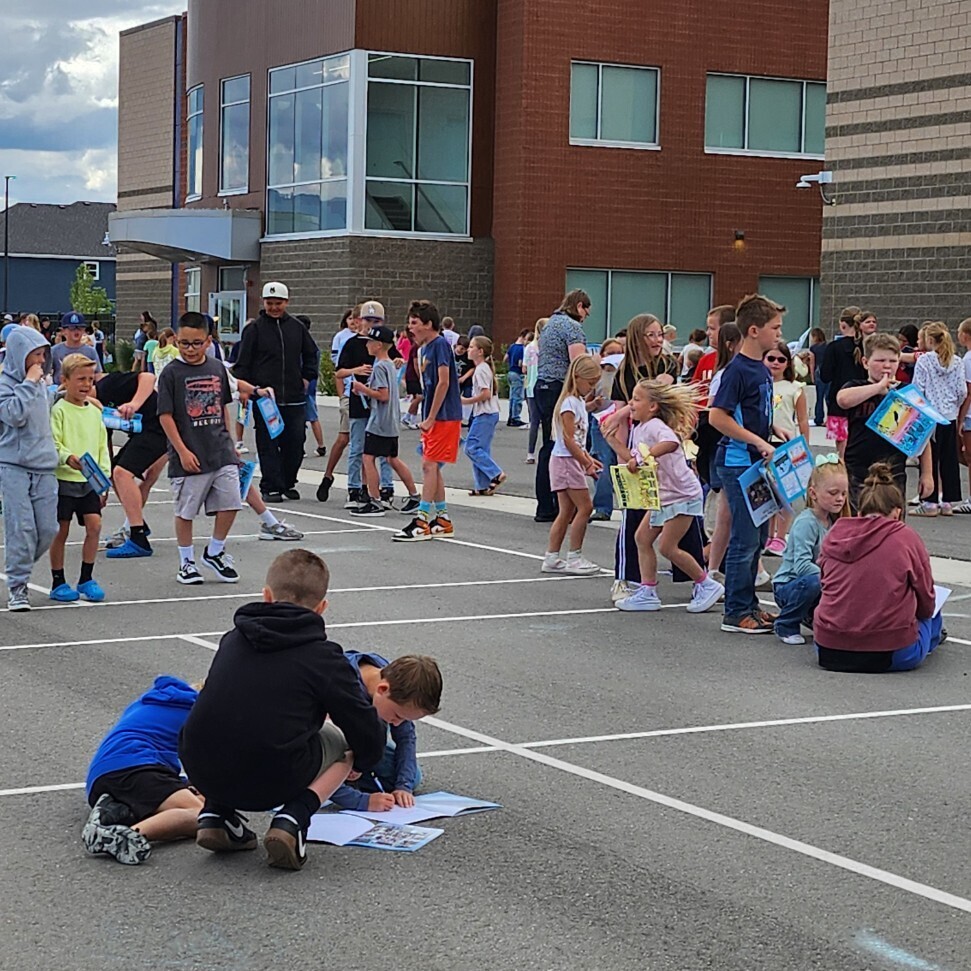 An outdoor shot of elementary school students gathered on an asphalt playground with white painted lines in front of a modern brown and brick school building. Several children are walking around holding blue folders, while two boys in the foreground crouch on the ground to write or sign yearbooks laid out on the pavement.