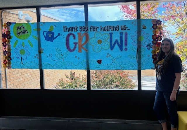 School counselor Mrs. Smith stands smiling next to a large, vibrant blue banner hanging in front of a window. The banner is decorated with flowers, a sun, and a watering can, with large colorful text that reads, "Mrs. Smith, thank you for helping us... GROW!" The background shows an outdoor courtyard with trees.