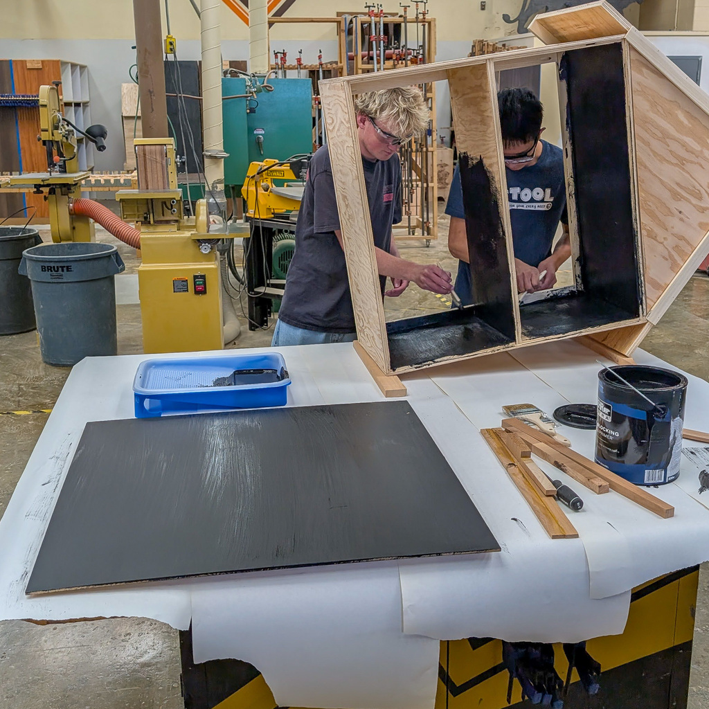 Two students are painting a tiny library in a wood shop