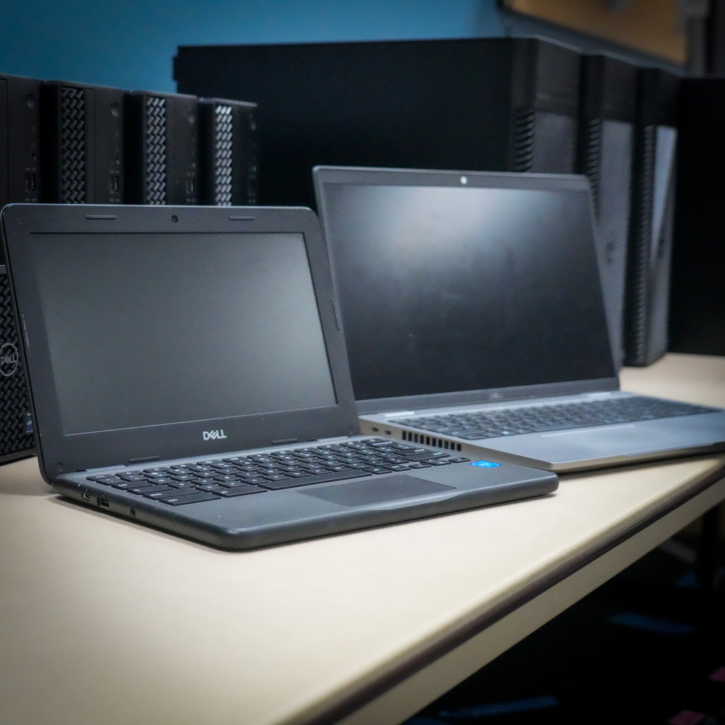 Two laptops are open on a desk with multiple computer towers behind 
