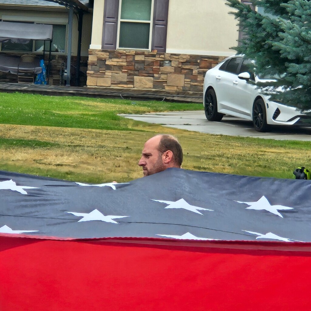 Lomond View Elementary custodian, Louis Child, carrying a massive American flag during a community parade in Pleasant View, Utah. Only his head and shoulders are visible above the large, vibrant flag.