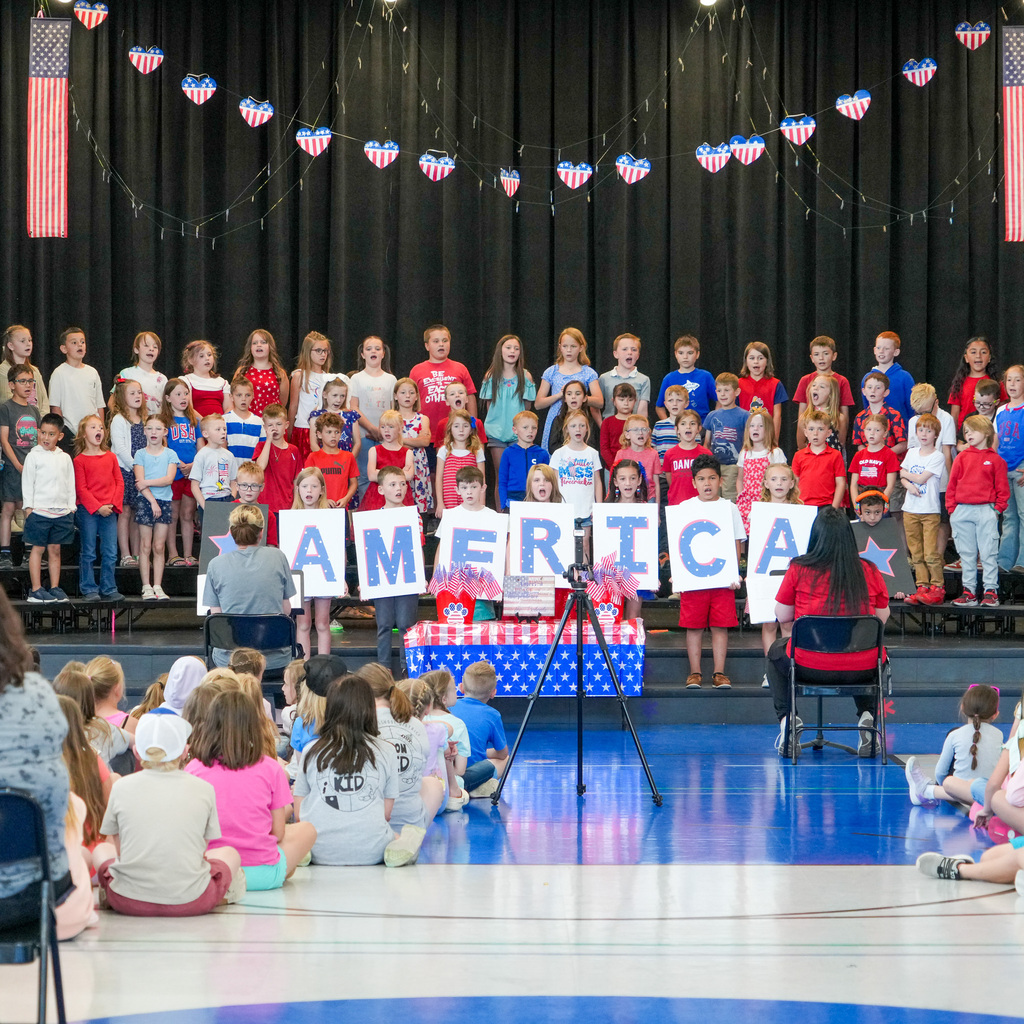 A group of students singing on a stage in a variety of red, white, and blue holding posters that spell out America