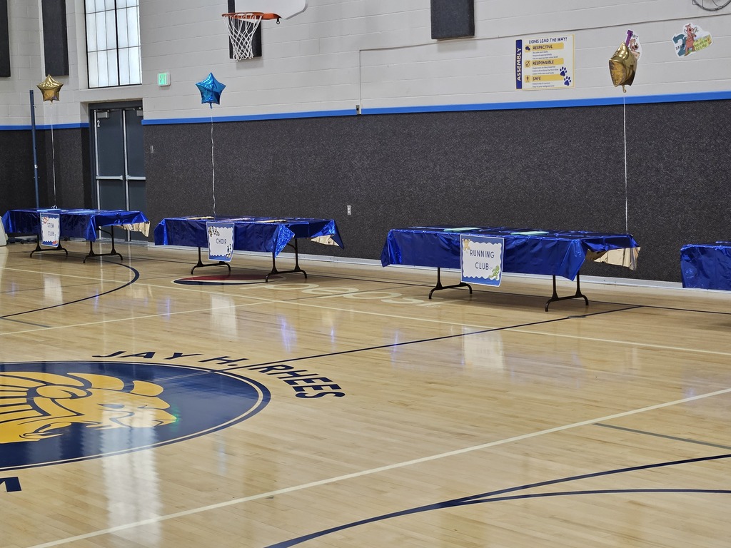 Display tables in a gymnasium feature labeled signs for student extracurricular activities, including STEM Club, Choir, and Running Club.