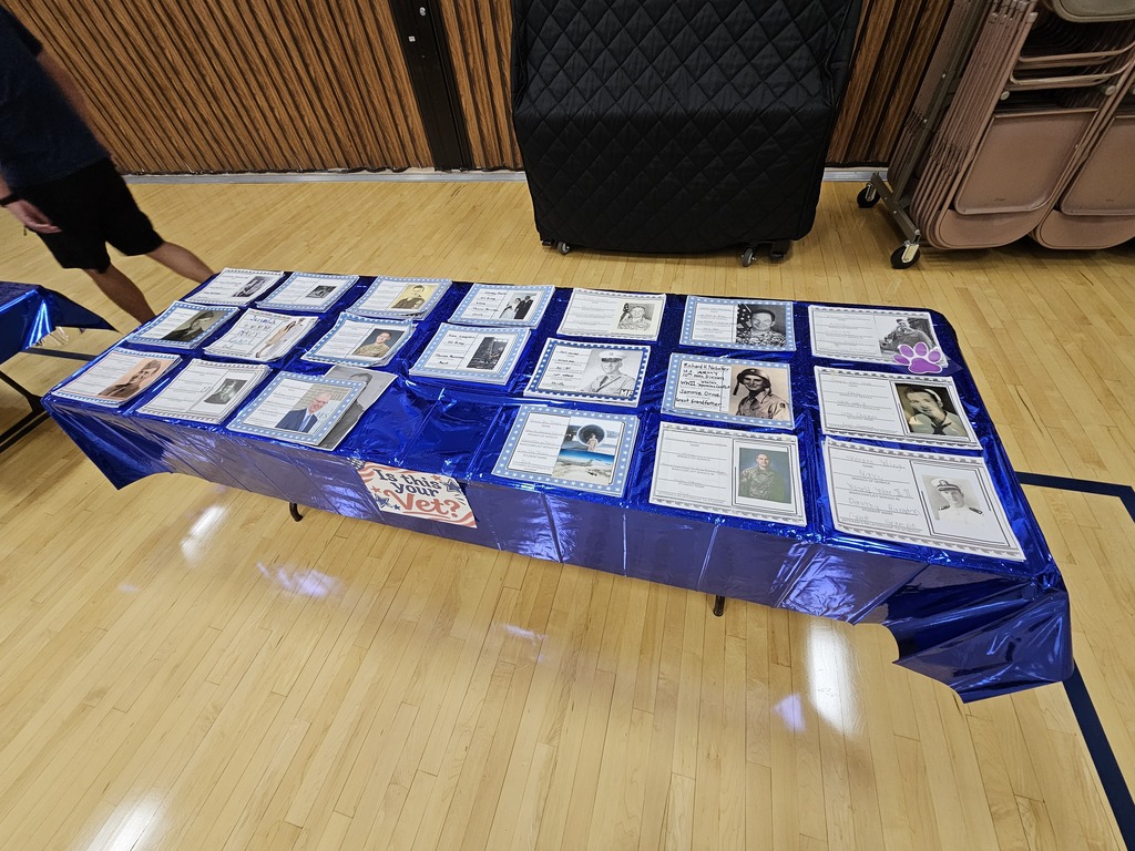 A display table covered with a blue metallic table runner presents framed photos and sheets honoring local military veterans in the school gymnasium.