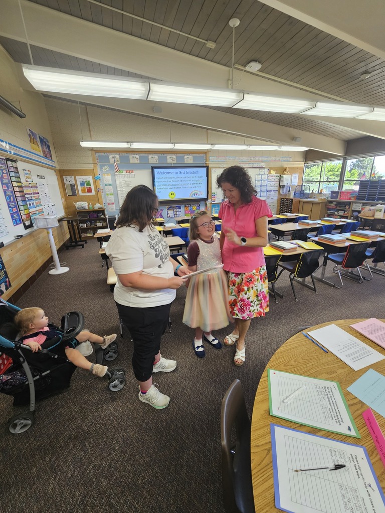 A third-grade teacher smiles and gestures warmly as she talks with a student and parent holding classroom paperwork inside a decorated classroom.
