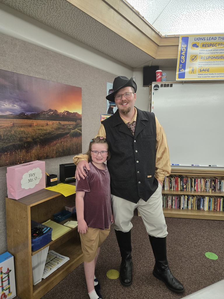 A fifth-grade teacher in a Musketeer costume and black hat smiles with his arm around a young student wearing glasses in a classroom.