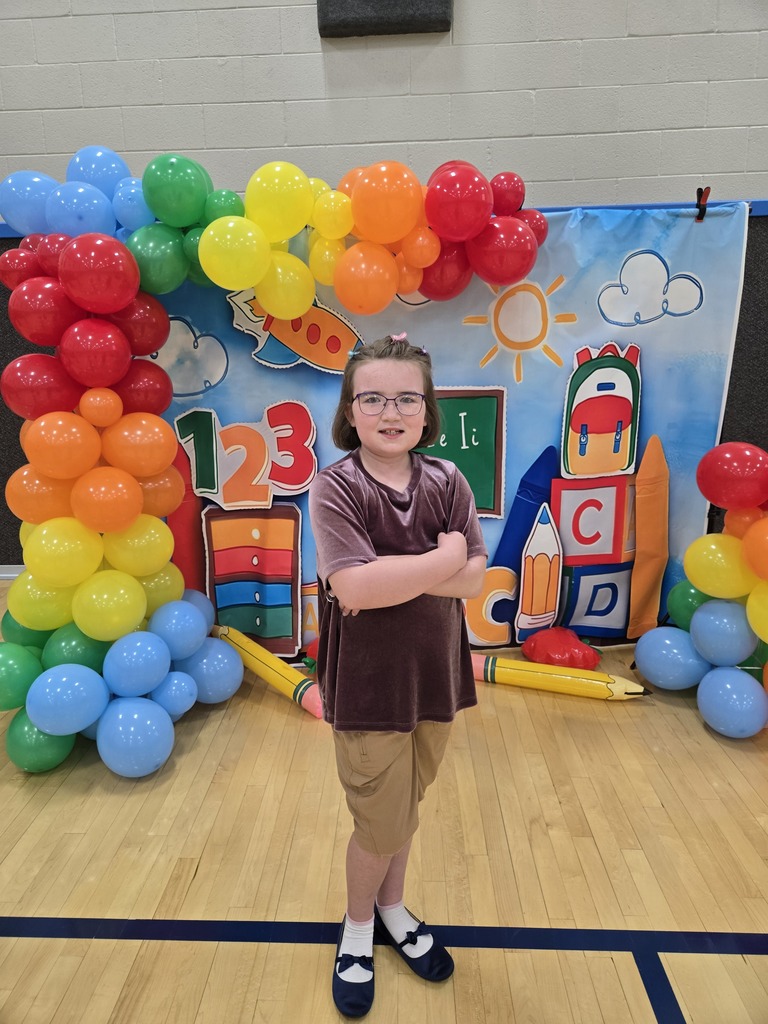 A student wearing glasses and a purple shirt poses with arms crossed in front of a colorful Back-to-School photo backdrop framed by a bright balloon arch.