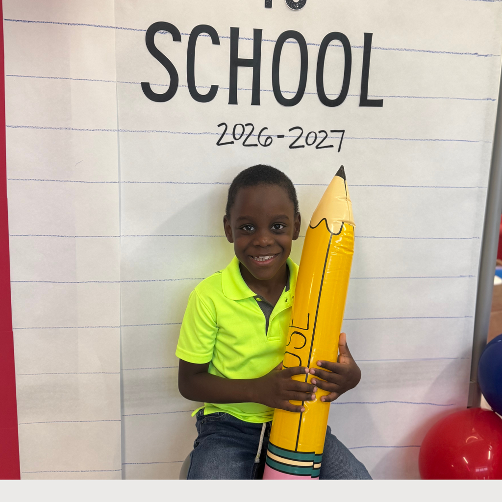 Student in front of a Back to School Sign with balloon arch holding a OSE large pencil. Theme red, white, and blue.