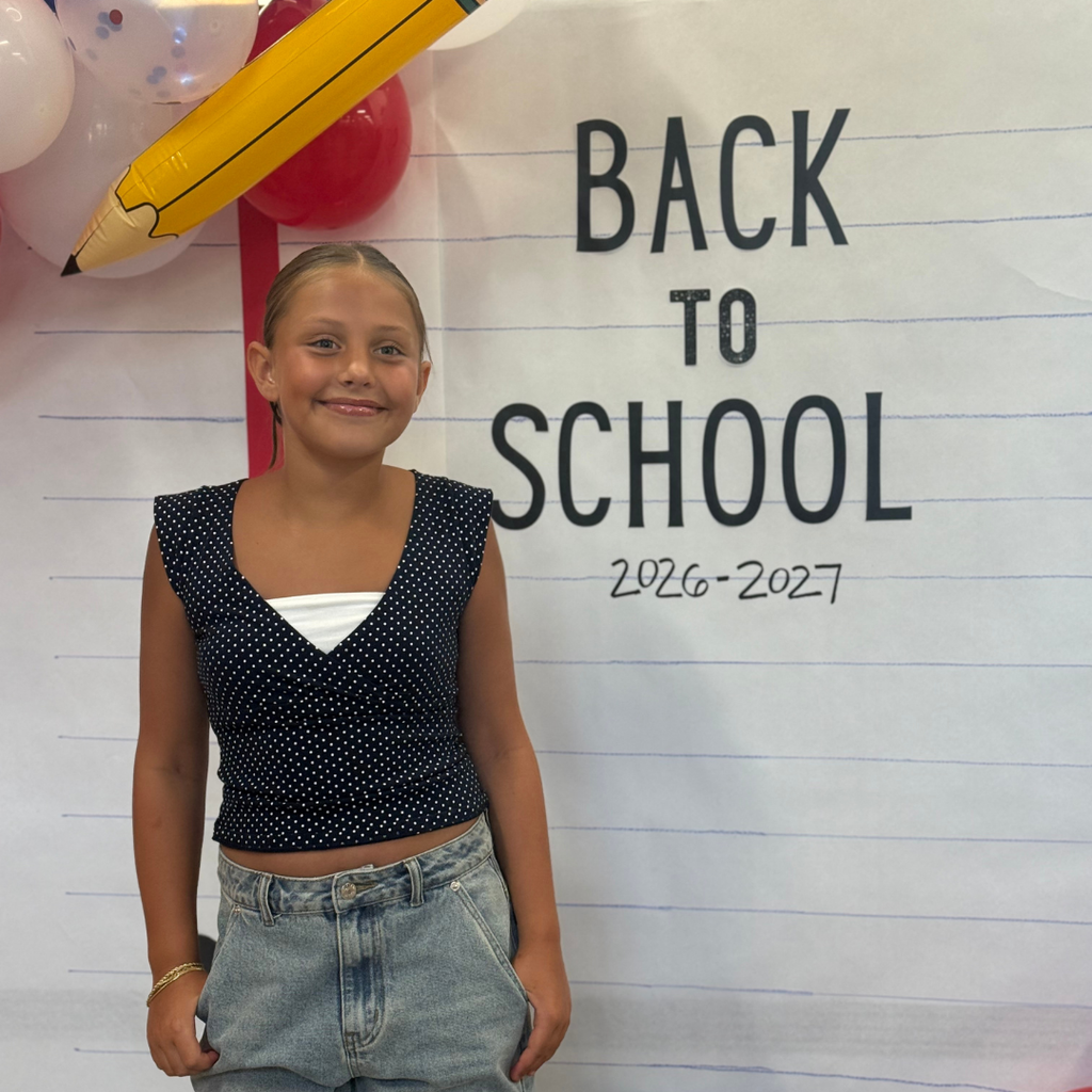 Student in front of a Back to School Sign with balloon arch holding a OSE large pencil. Theme red, white, and blue.