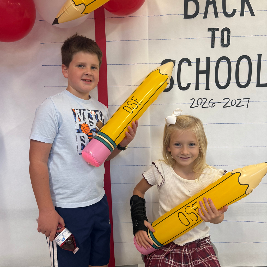 Students in front of a Back to School Sign with balloon arch holding a OSE large pencil. Theme red, white, and blue.