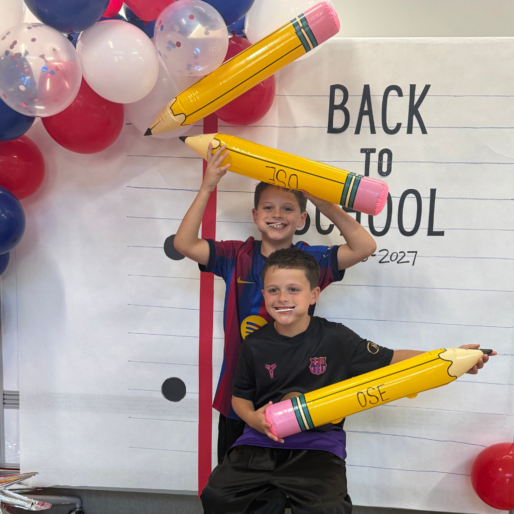 Students in front of a Back to School Sign with balloon arch holding a OSE large pencil. Theme red, white, and blue.
