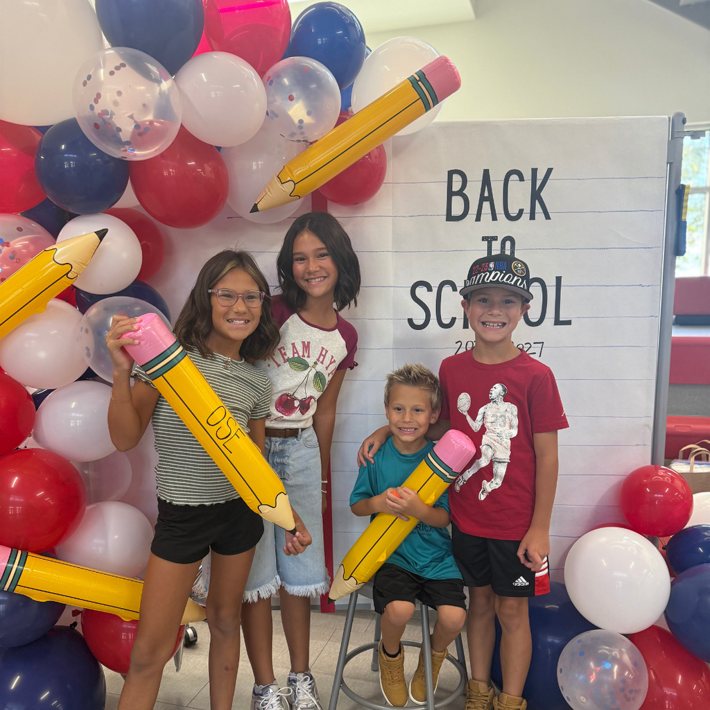 Students in front of a Back to School Sign with balloon arch holding a OSE large pencil. Theme red, white, and blue.