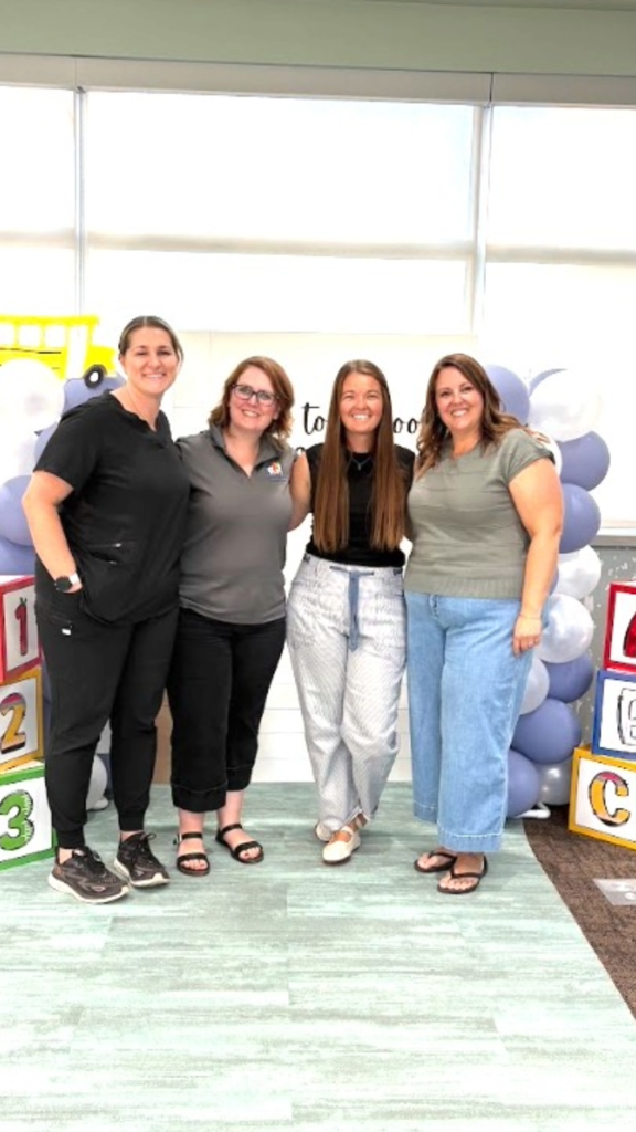 Four women standing shoulder-to-shoulder smiling for a photo in front of a backdrop showing the text "Back to School". The setup is flanked by a balloon column topped with a school bus figure on the left, and stacked 1, 2, 3 and A, B, C toy blocks on the outer edges.